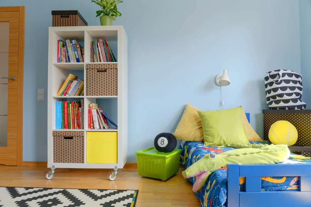 Cuarto de hijos con cama azul decorada con una estantería blanca con libros y cajas.