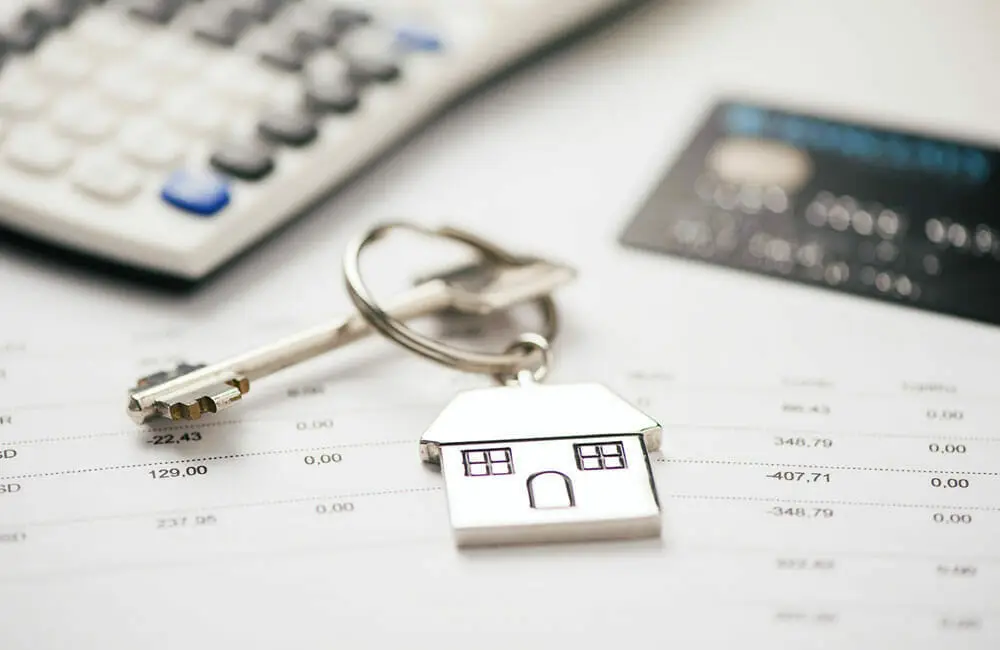 Llavero en forma de casa con llave, tarjeta de crédito y calculadora sobre hojas de papel con cifras.