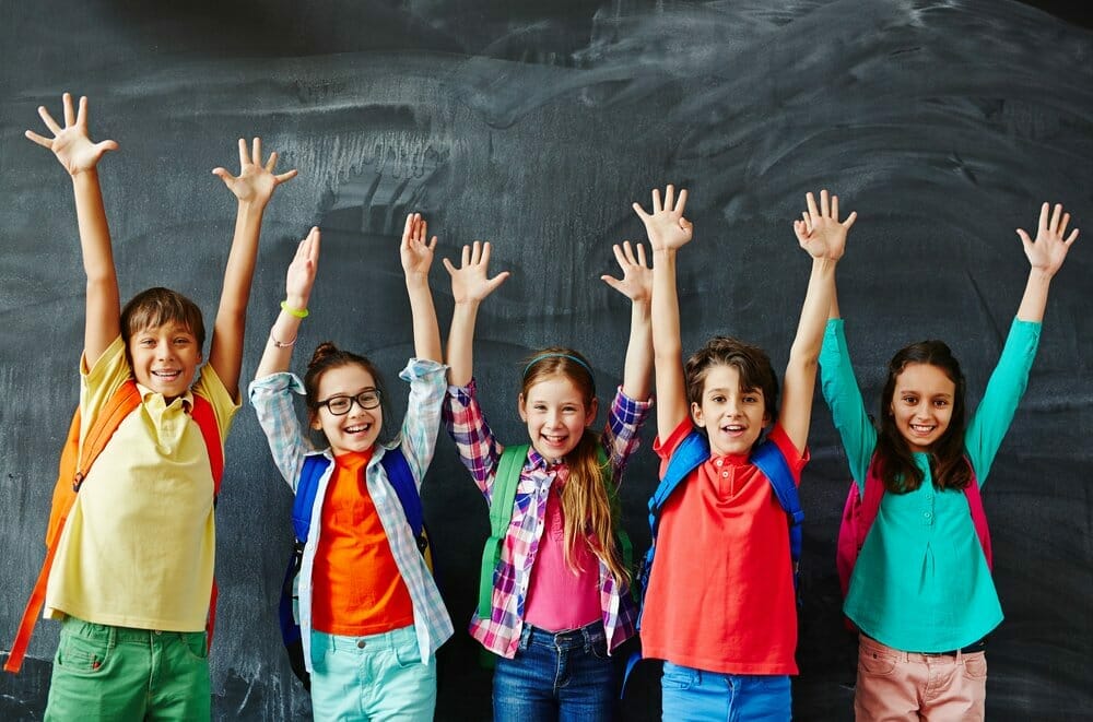 3 chicas y 2 chicos con las manos levantadas y de espalda a la pizarra en la clase del colegio.