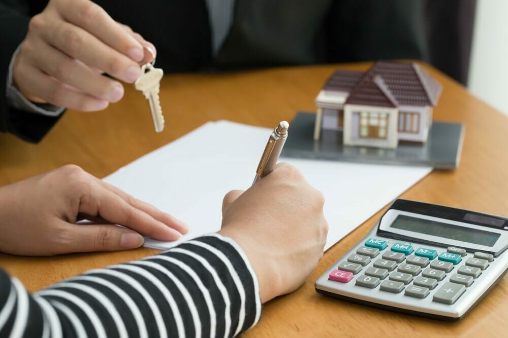 Mujer firma un contrato para comprar una casa mientras que un agente de bienes raíces le ofrece la llave de su nueva casa.