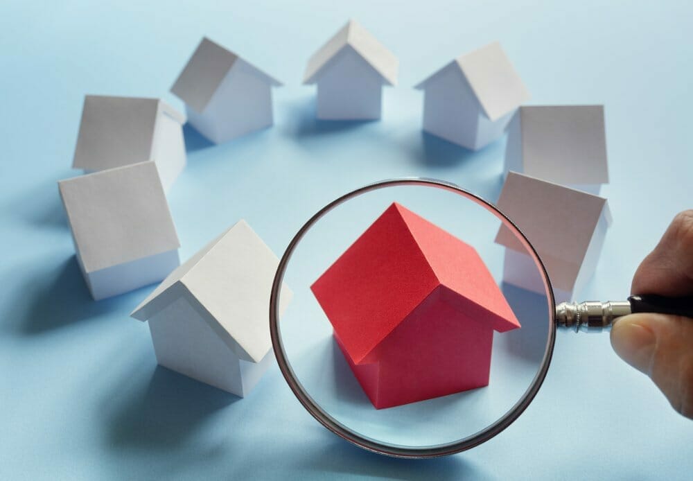 Ocho pequeñas casas blancas y una roja sobre fondo azul y una lupa amplificando la casa roja.